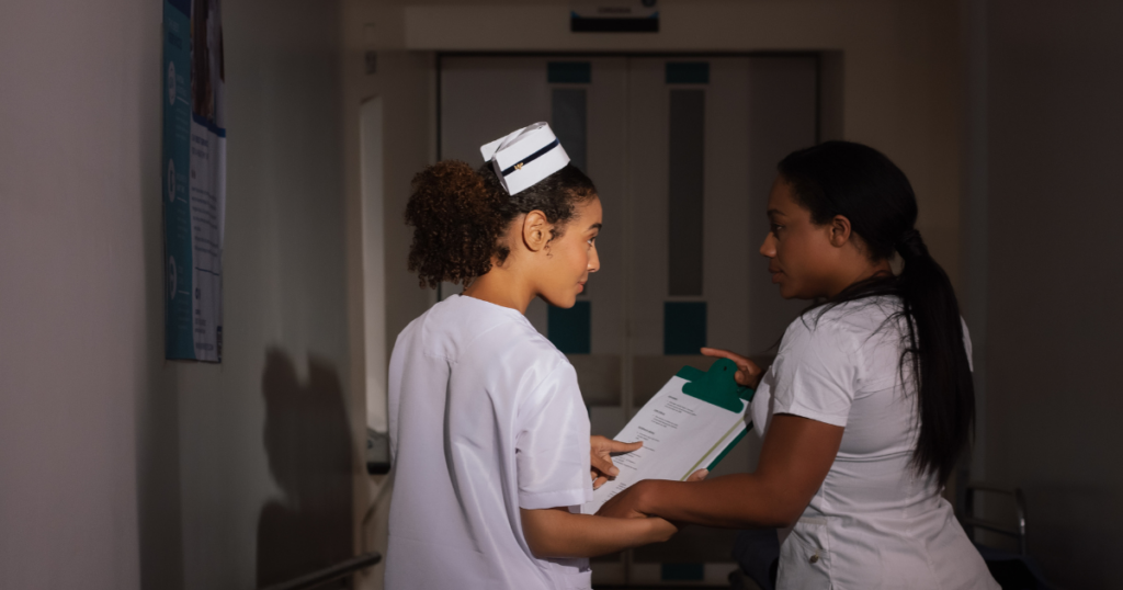 Image of 2 nurses standing, discussing a case in a low lit room. The Weekend Shift