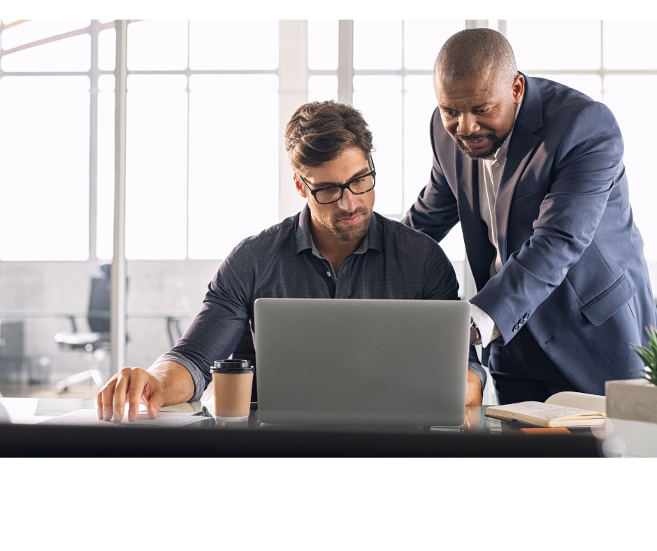 Image of a man sitting at his computer with another man helping him. Leadership Confidence: Why Leading Without Being the Expert Feels So Uncomfortable and What Leadership Actually Requires