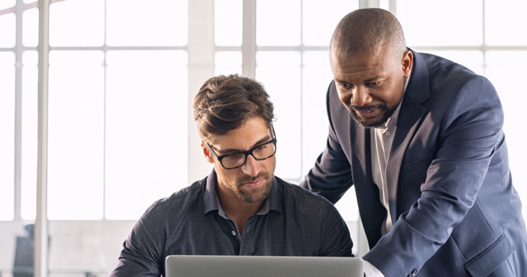 Image of a man sitting at his computer with another man helping him. Leadership Confidence: Why Leading Without Being the Expert Feels So Uncomfortable and What Leadership Actually Requires