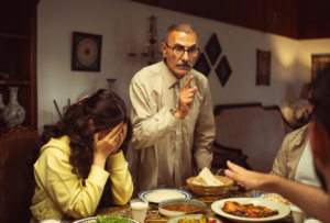 Image of a man standing sternly in rebuke at the dinner table with daughter beside him with her head in her hands in frustration. Family estrangement