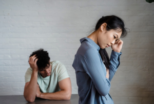 Woman standing in forefront holding her head down in regret, while man sitting in background holding his head down in regret. Financial secret