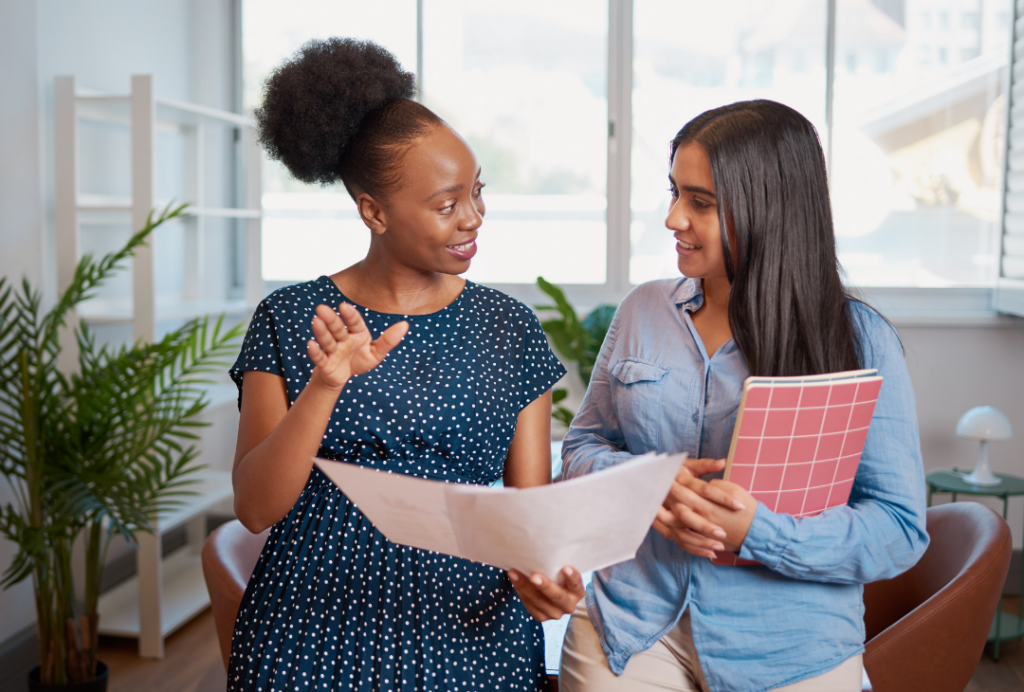 Image of two women interacting while one explains what's on a sheet of paper to the other. Mentorship