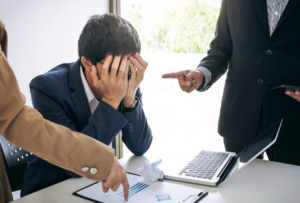 Image of man sitting at his computer overwhelmed by two people pointing in demand at him. Overbearing behavior