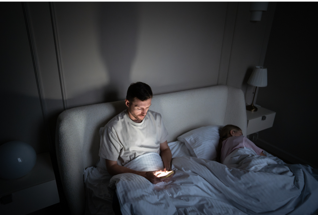 Image of a man sitting up in bed in a dark room, using his phone, while his partner is asleep beside him. Screen overuse