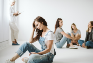 Image of a woman in the foreground looking sad while a group of women hang are talking casually at the back. Social exclusion